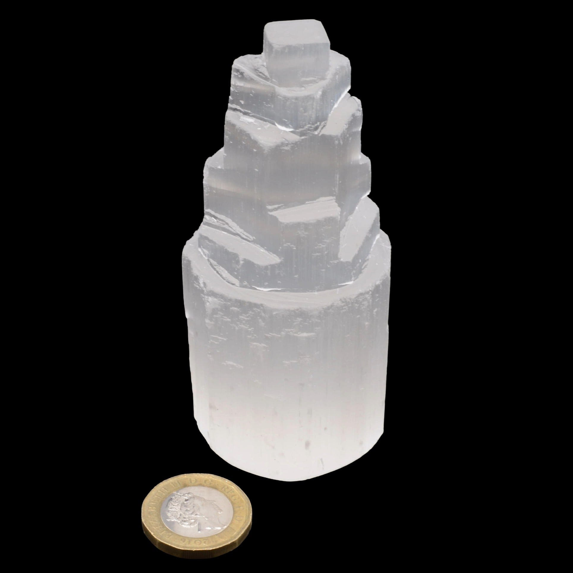 A translucent white selenite crystal tower with a stepped, fractal-like structure, shown on a solid black background with a gold and silver coin below for scale.