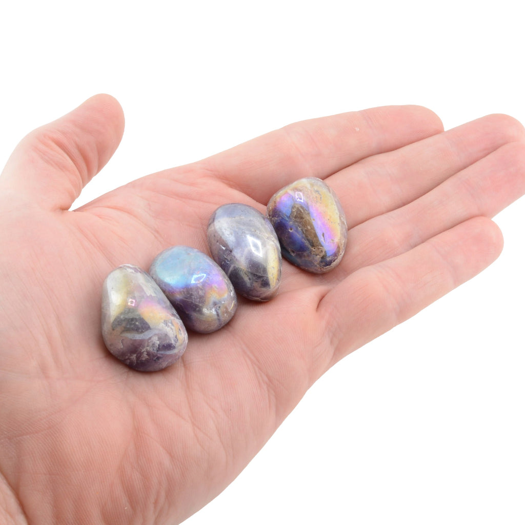 Four polished iridescent tumbled  aura amethyst gemstones in purple, blue, and gold hues resting on an open human hand against a plain white background.